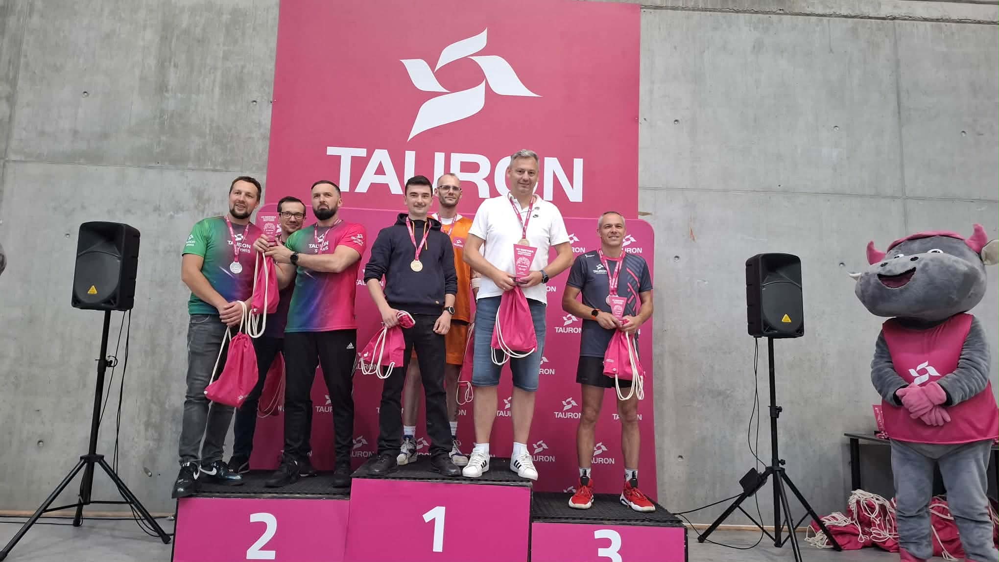 Zwycięzcy stoją na podium. W tle logo TAURON i maskotka byczek.