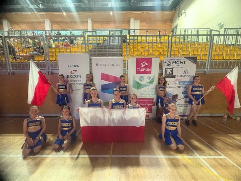 Cały zespół pozuje na hali. Laureatki trzymają flagi Polski. W tle loga sponsorów.