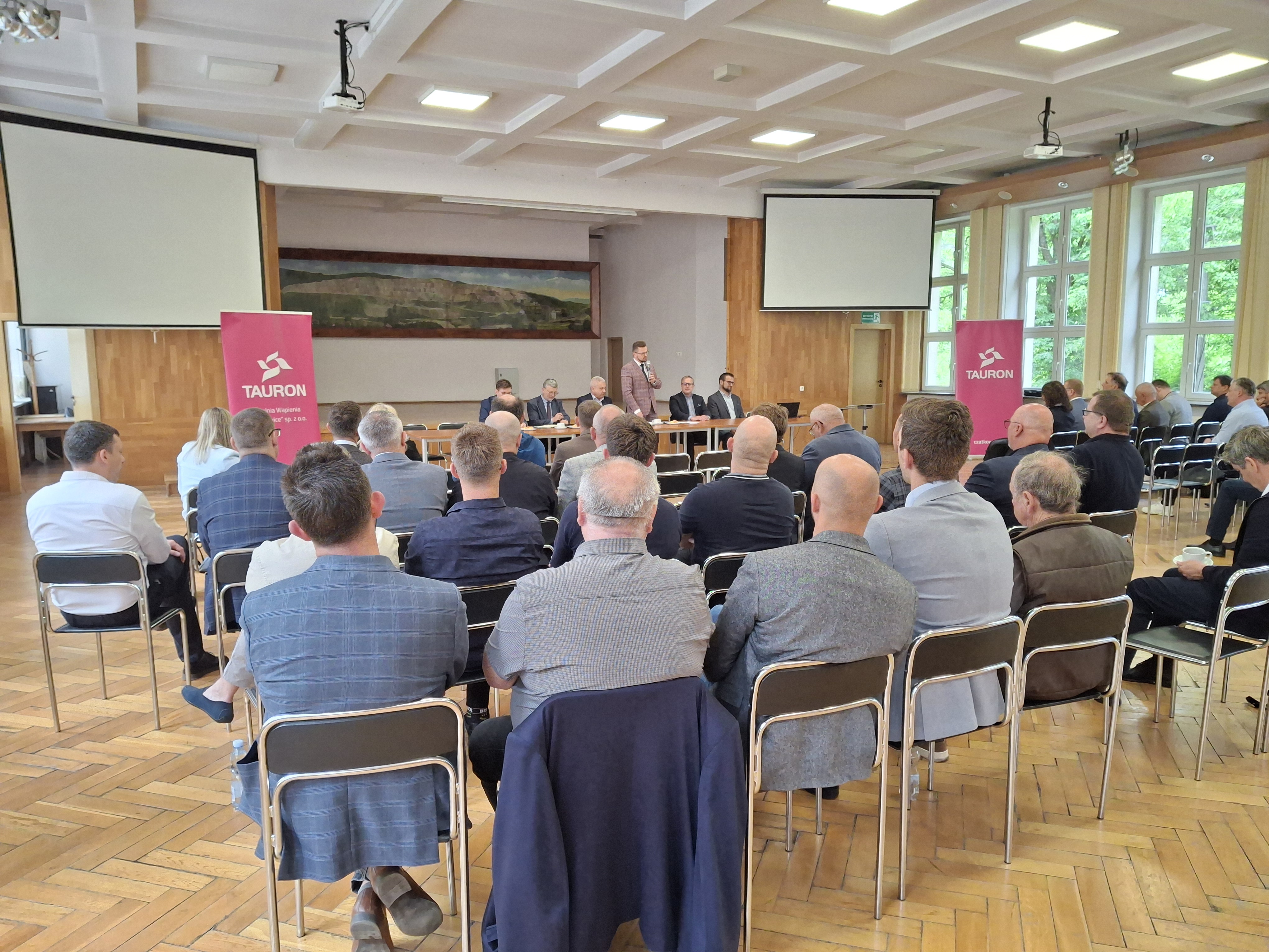 Uczestnicy spotkania siedzą na auli i słuchają wystąpienia. W tle logo TAURON