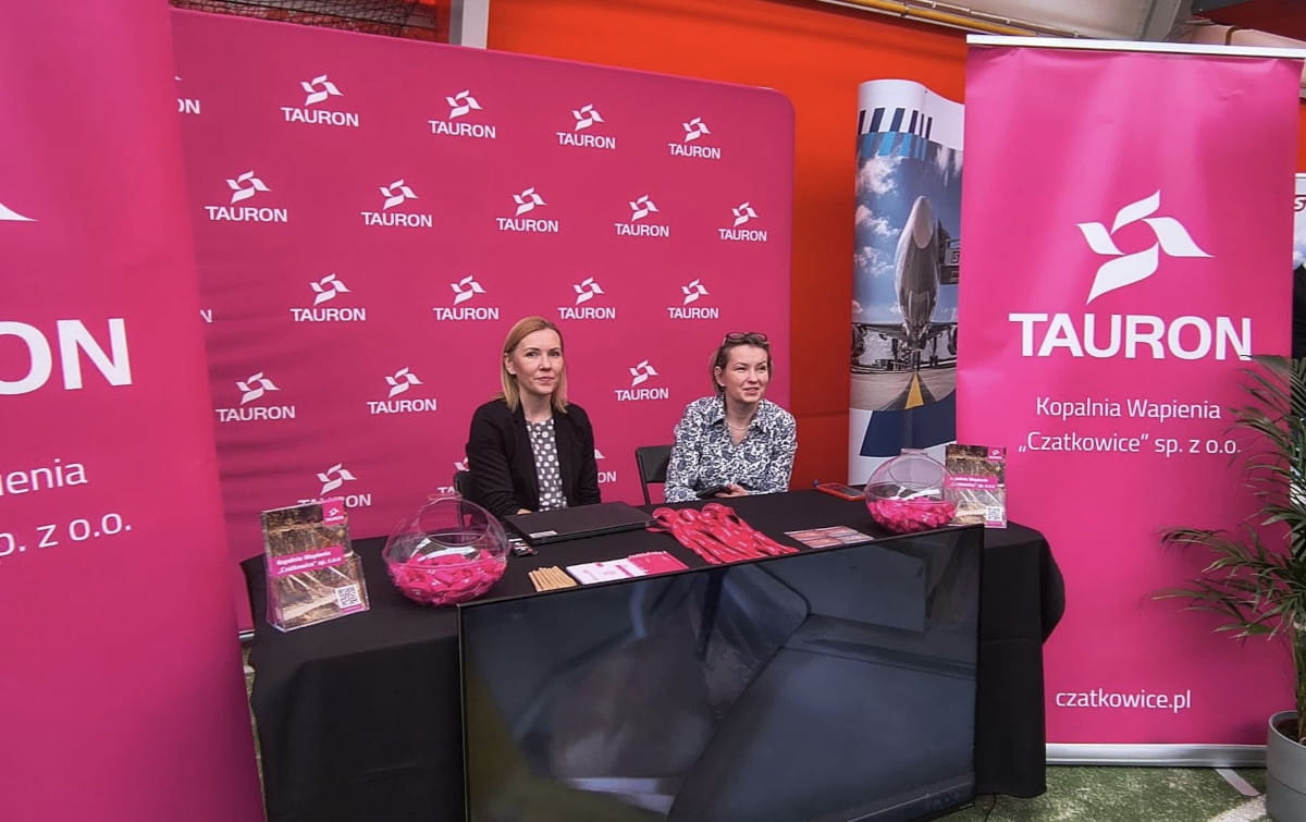 Przedstawiciele Kopalni Wapienia “Czatkowice” sp. z o.o. siedzą przy stoisku Spółki z ulotkami dotyczącymi praktyk i gadżetami. W tle ścianka i rollupy Grupy TAURON.