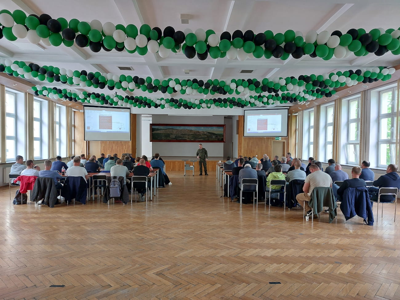 Szkolenie dla pracowników w sali konferencyjnej – uczestnicy siedzą przy stołach, a prowadzący stoi przed nimi; sufit ozdobiony girlandami z zielonych, białych i czarnych balonów.