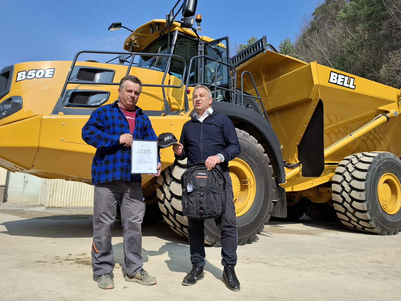 Pracownik Kopalni Wapienia "Czatkowice" sp. z o.o. w koszuli w kratkę z dyplomem ukończenia kursu kierowania wozidłem Bell B50e z przedstawicielem firmy Kiesel Poland. W tle wozidło Bell B50e